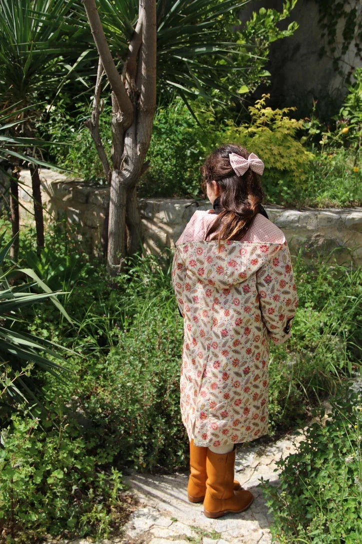 Child in floral print raincoat with bow hair clip and brown boots standing in lush garden