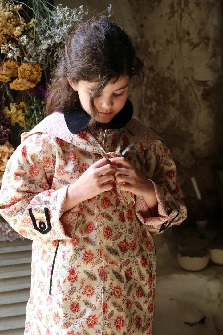 Young girl wearing a floral patterned coat with black collar, standing indoors by dried flowers