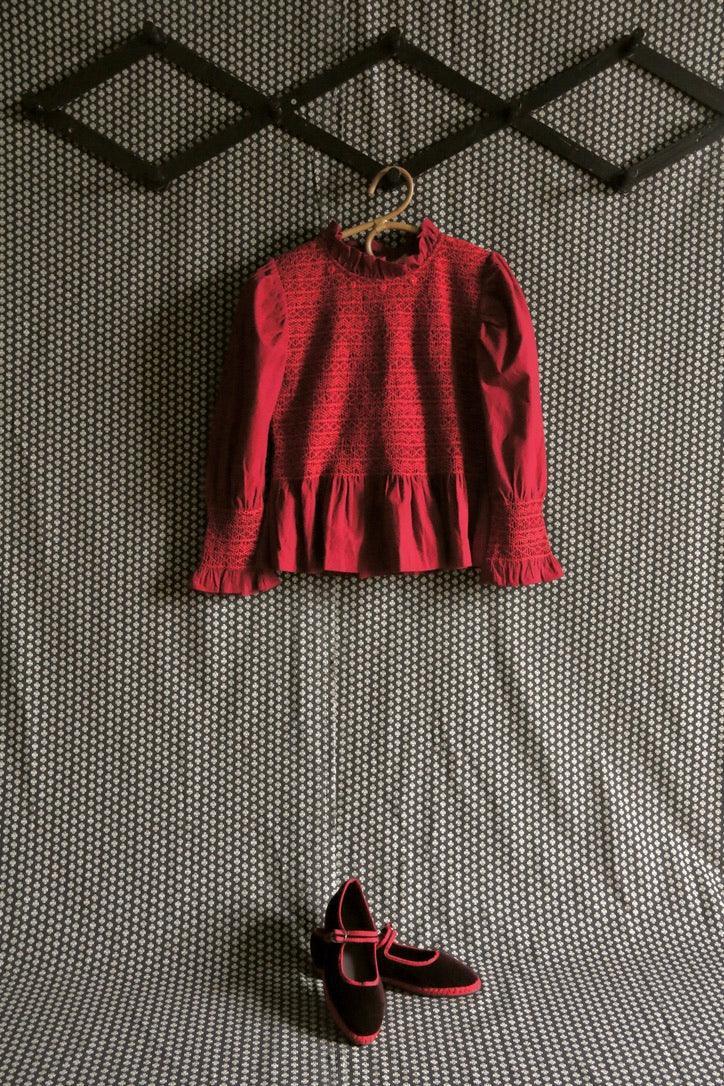 Red smocked long sleeve girls blouse and matching Mary Jane shoes on patterned background