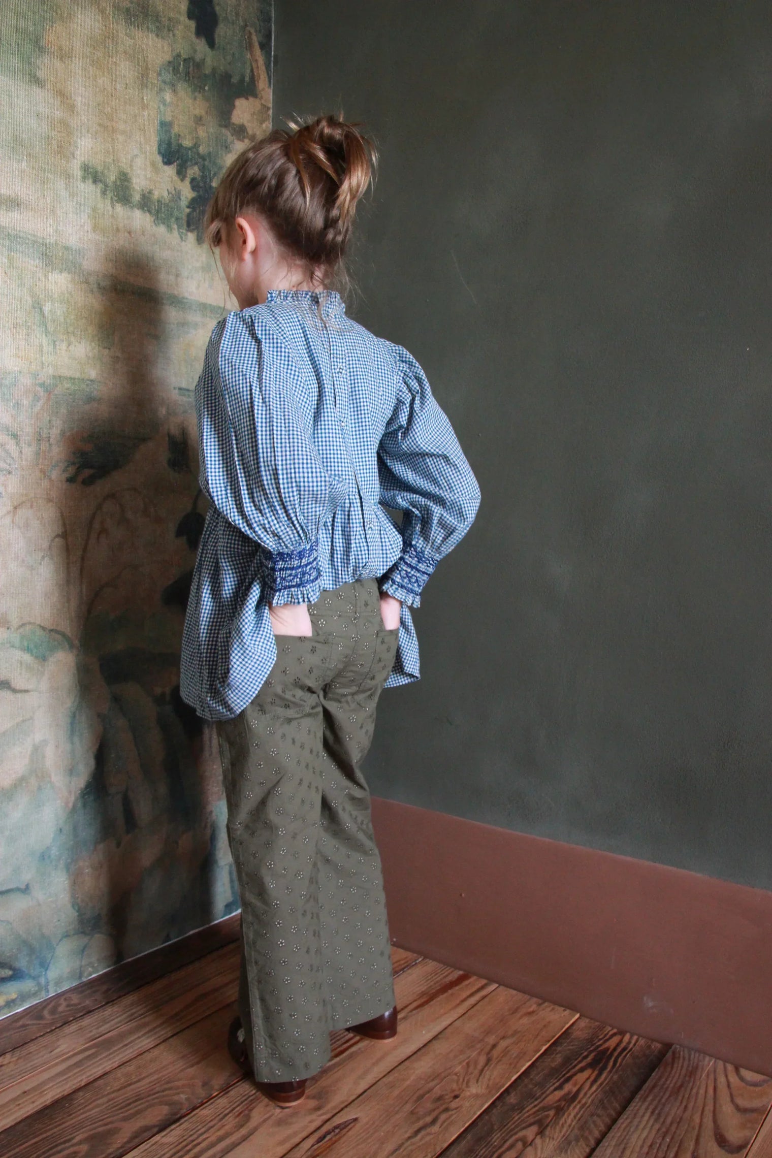 Girl in blue gingham blouse and olive embroidered pants standing on wooden floor indoors