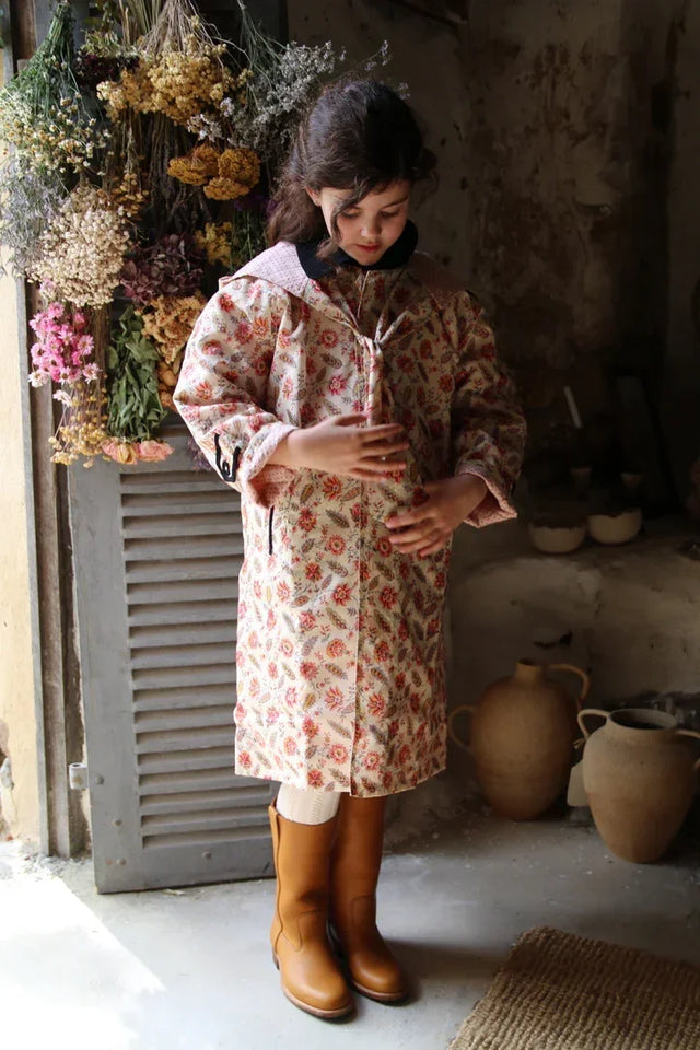 Girl in floral print long coat, tan boots, standing indoors by hanging dried flowers