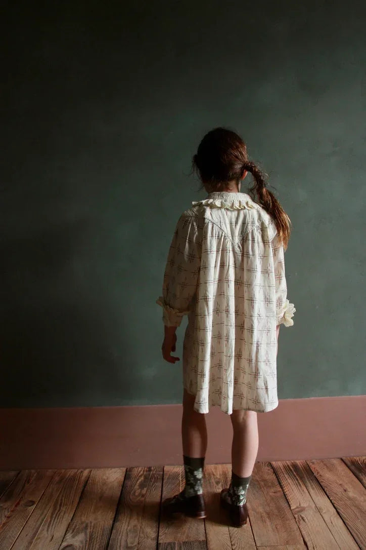 Child in loose printed dress with ruffled collar, standing on wooden floor indoors
