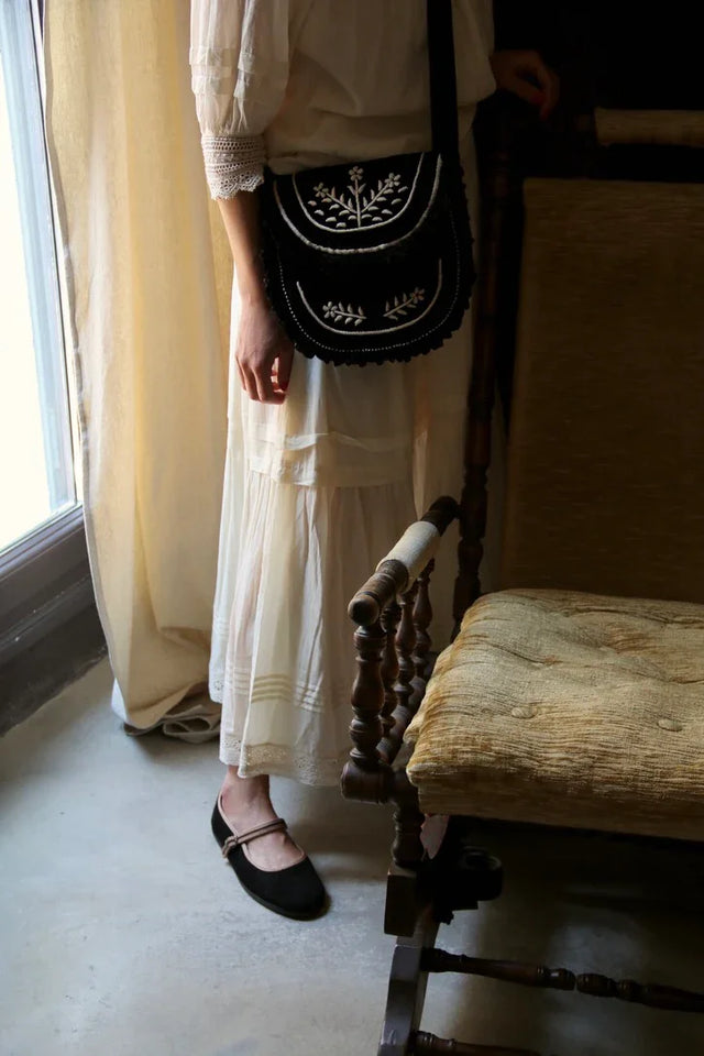 Woman in white dress with black embroidered shoulder bag and black flats standing indoors