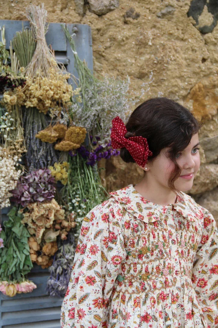 Girl in floral dress with red polka dot bow, standing by dried flower arrangement and rustic wall