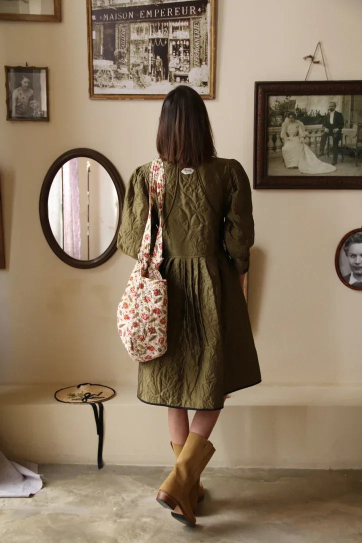Woman in quilted olive dress with floral tote bag and boots, vintage framed photos on wall