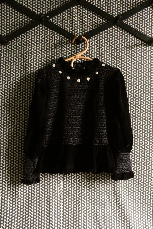 Black velvet long sleeve children's blouse with lace details and floral buttons, hanging on a patterned wall