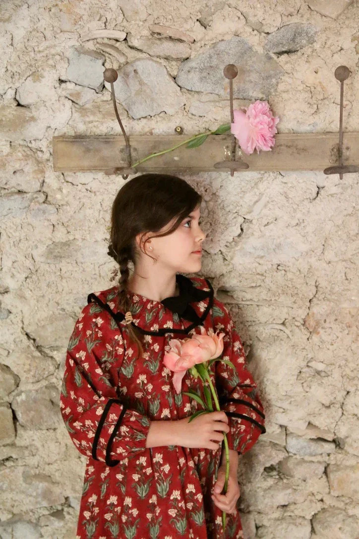 Girl in floral red dress holding pink flowers, standing by rustic stone wall
