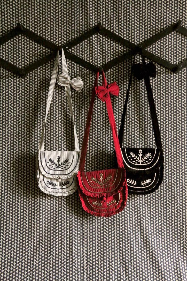 Three embroidered children's shoulder bags in striped, red, and black with bows, hung on patterned wall