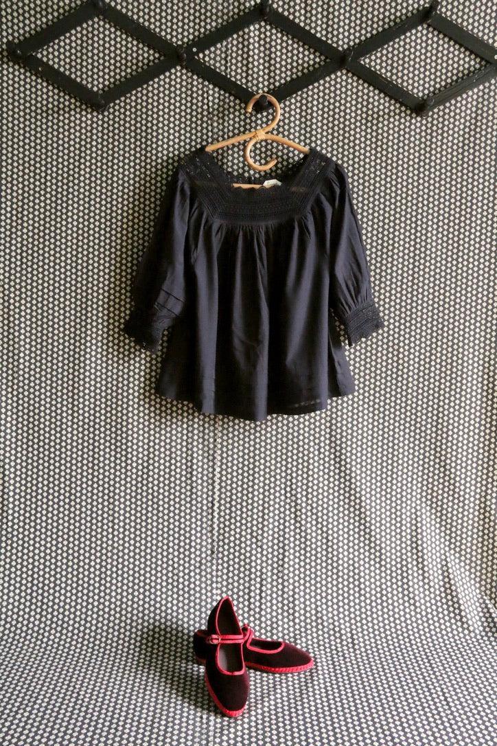 Black smocked girls blouse and black Mary Jane shoes with red trim on geometric background