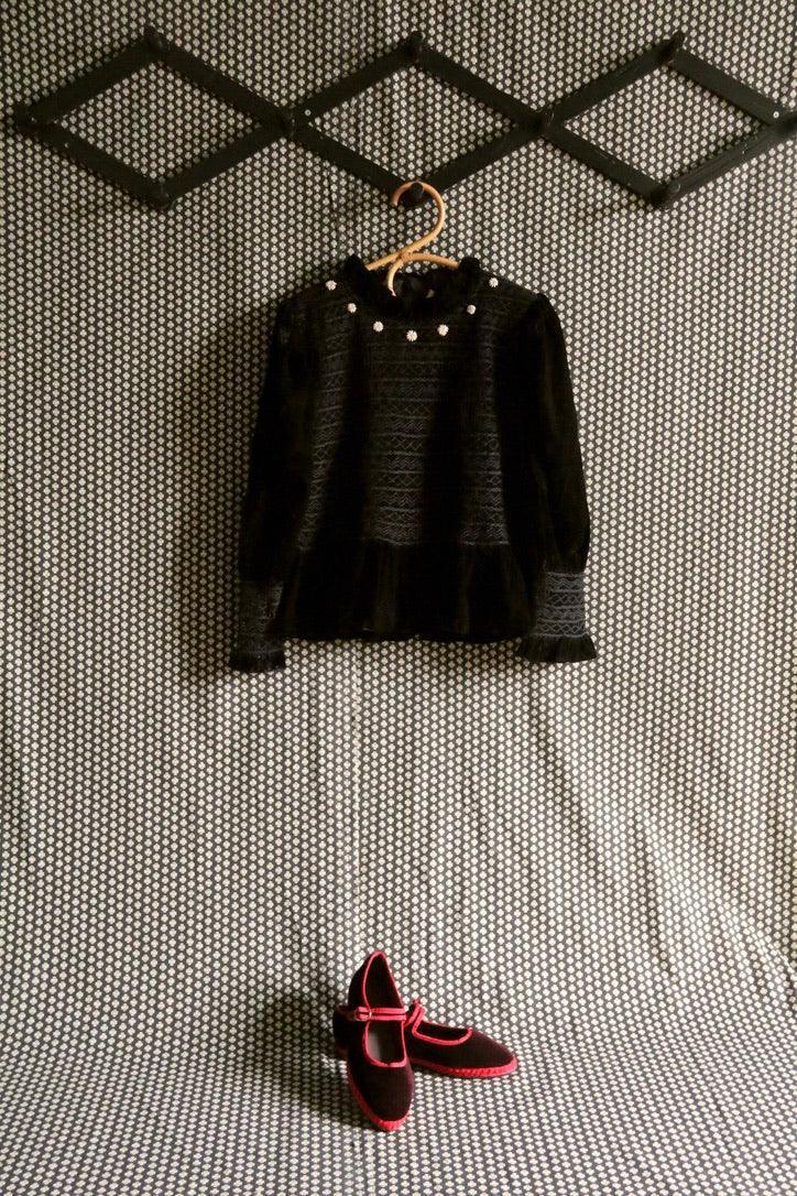 Black velvet children's blouse with pearl accents and black shoes with red trim on patterned background