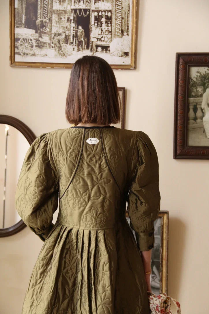Woman in olive green quilted dress with puff sleeves, standing in vintage style room