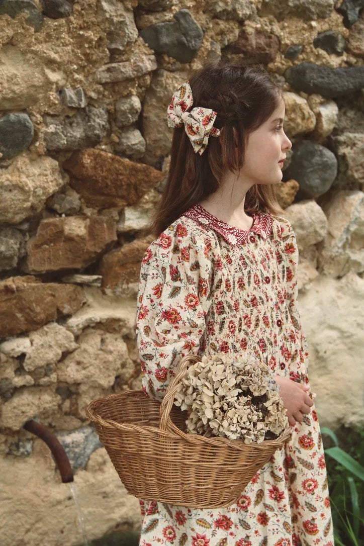 Girl in vintage floral dress with matching hair bow holding wicker basket of dried flowers, rustic stone wall background