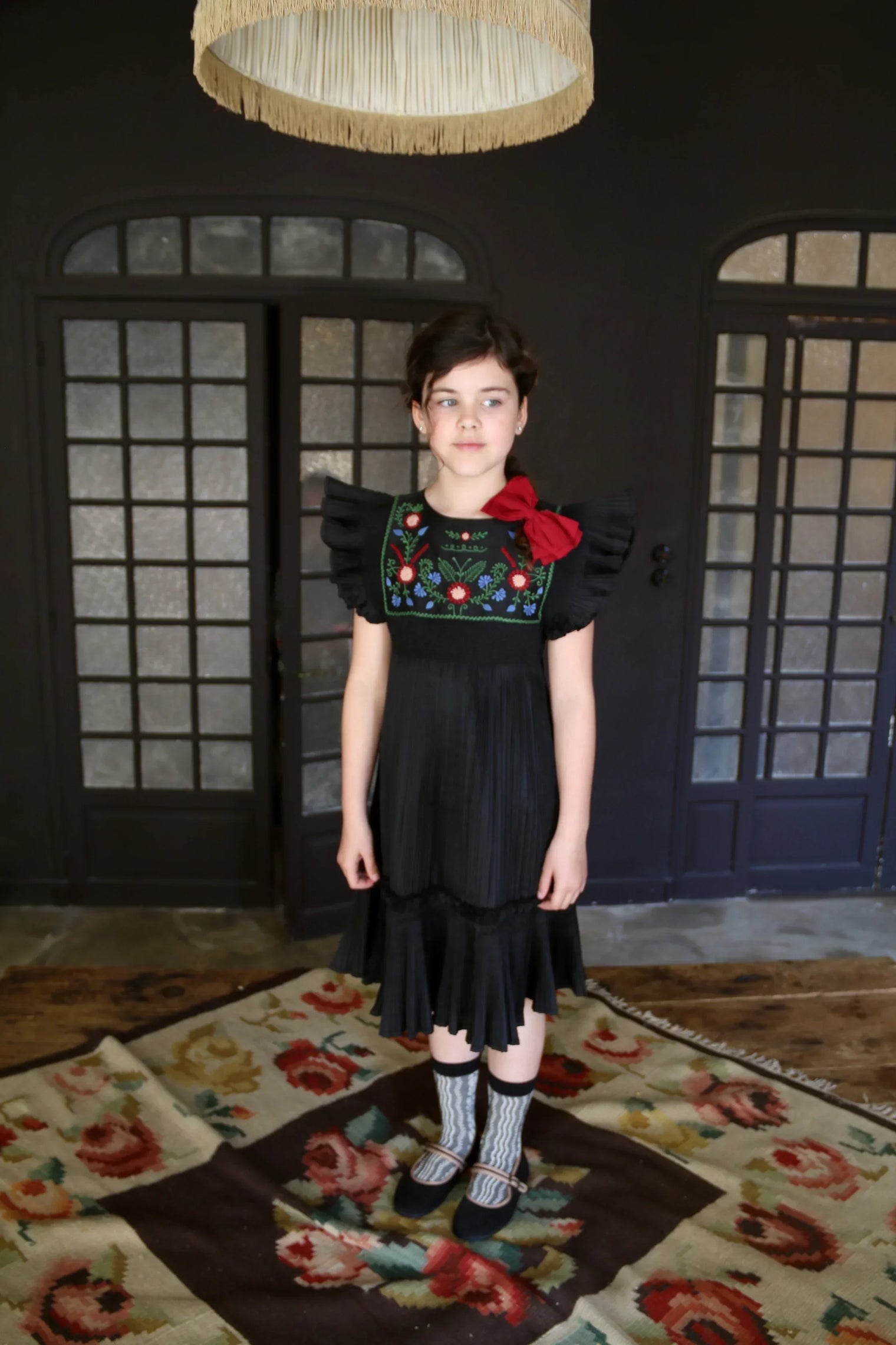 Girl wearing black dress with floral embroidery and ruffled sleeves, standing on floral rug indoors