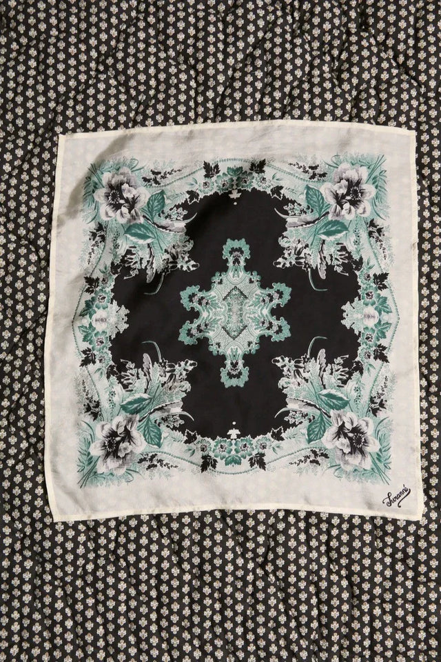 Square floral print scarf in green and black tones on patterned fabric background