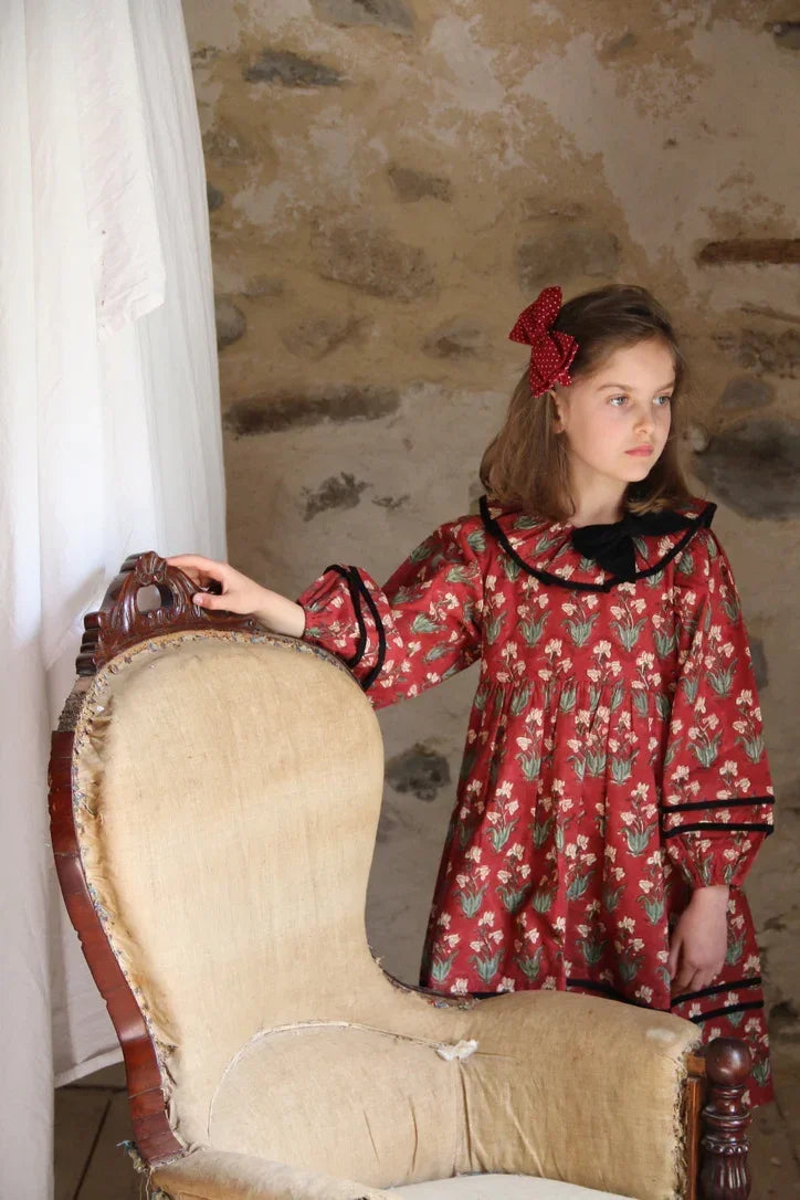 Girl in vintage floral print red dress with black trim and bow, standing by antique chair indoors