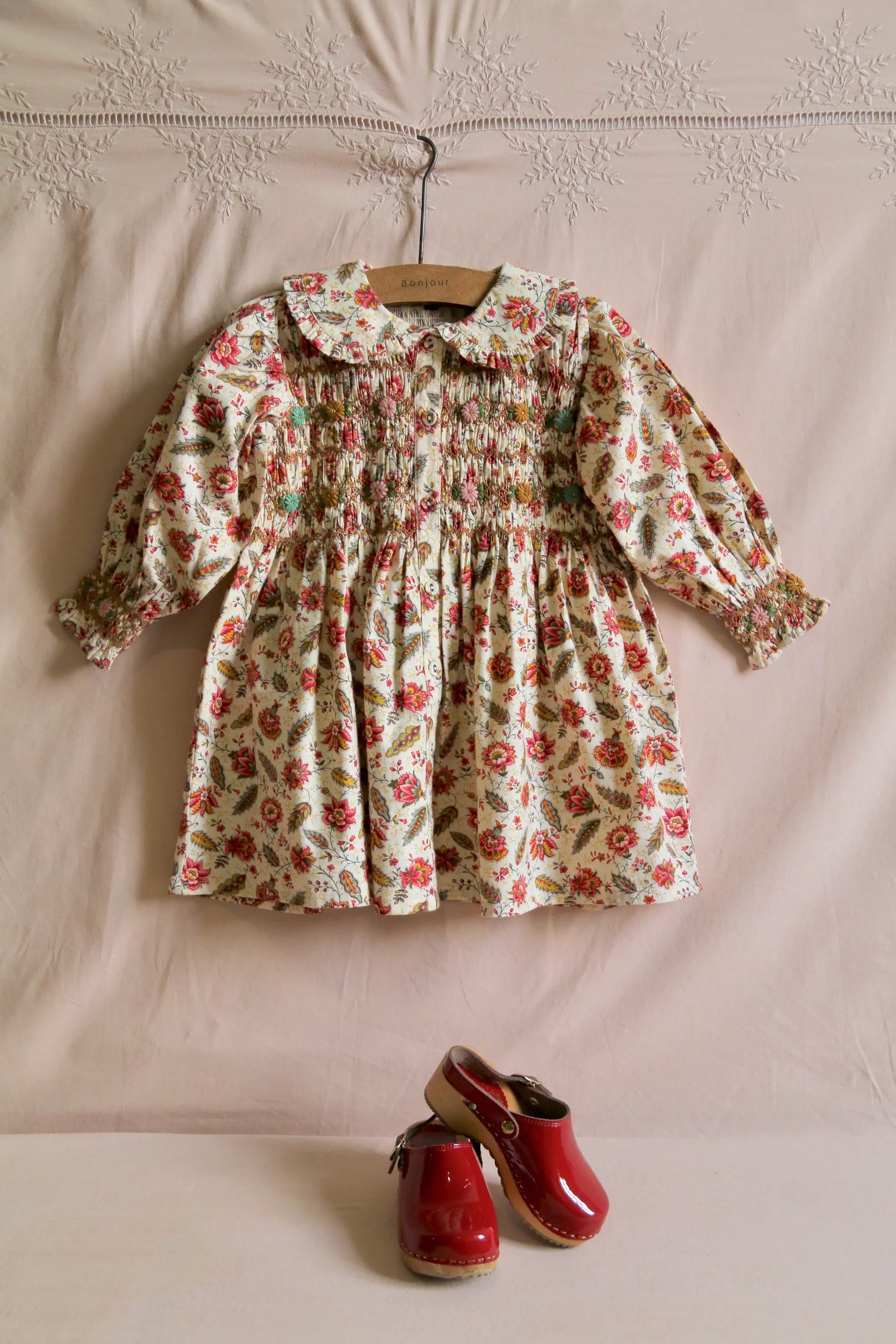 Floral smocked long sleeve dress for children with red wooden clogs on light background