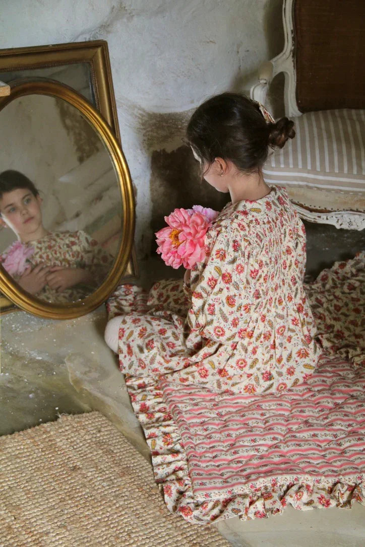 Child in vintage floral dress holding pink flowers sits on patterned cushion, reflected in mirror
