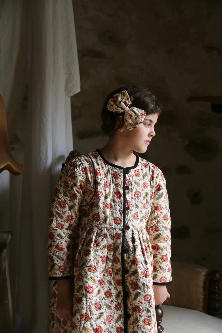 Girl wearing vintage floral quilted dress and matching headband, standing indoors by window