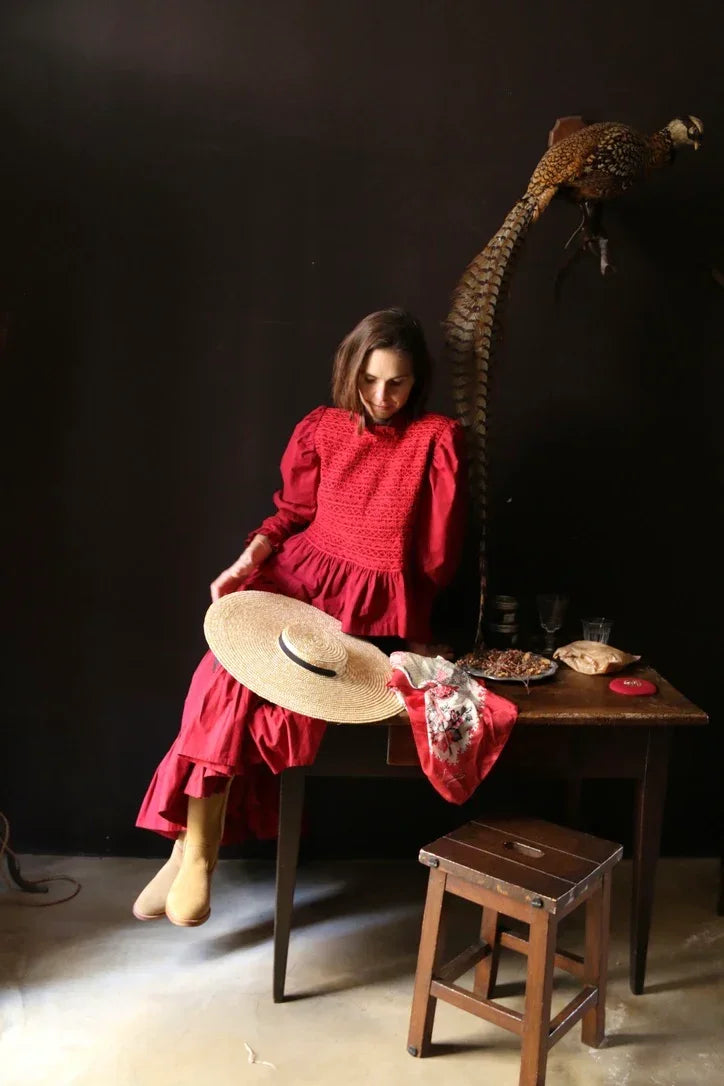 Woman in a long red dress with puff sleeves sitting by a table with a straw hat and scarf, vintage decor background