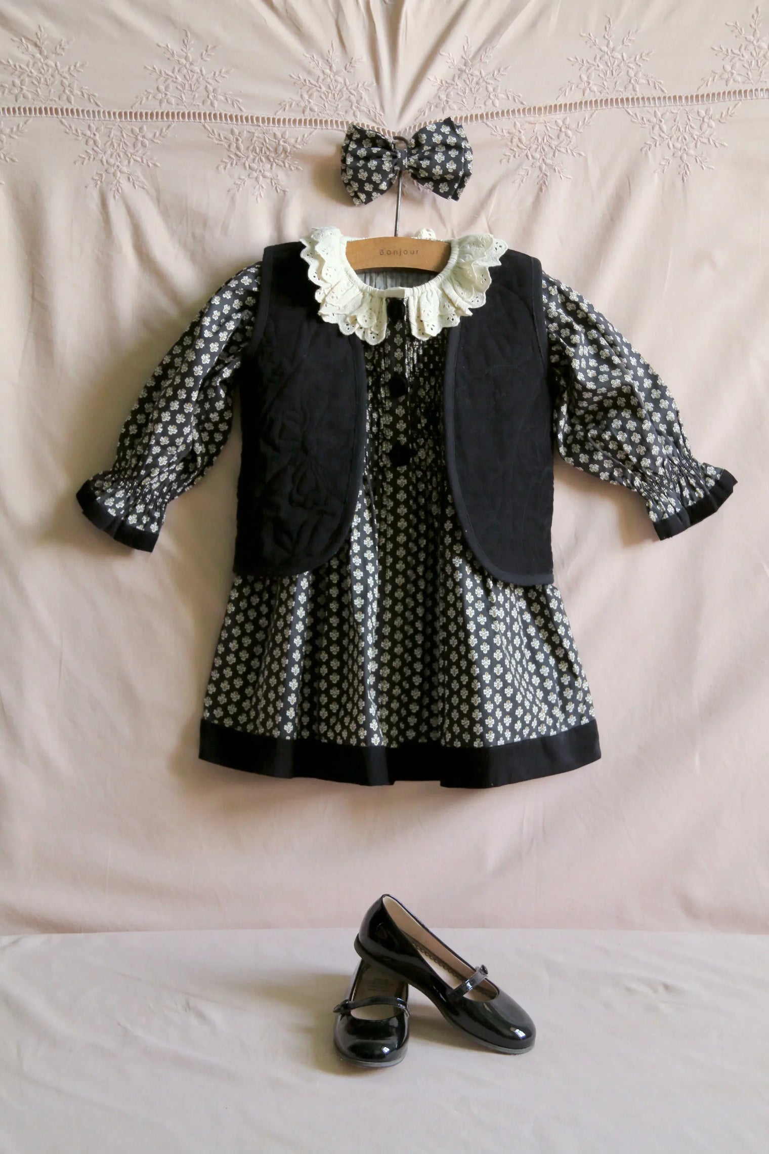 Children’s black floral dress with lace collar, matching vest, hair bow, and patent shoes.