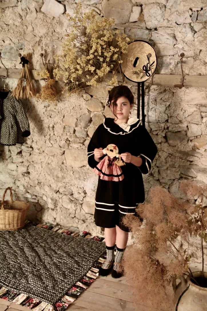 Girl in vintage childrenswear holding doll, standing by rustic stone wall with dried flowers.