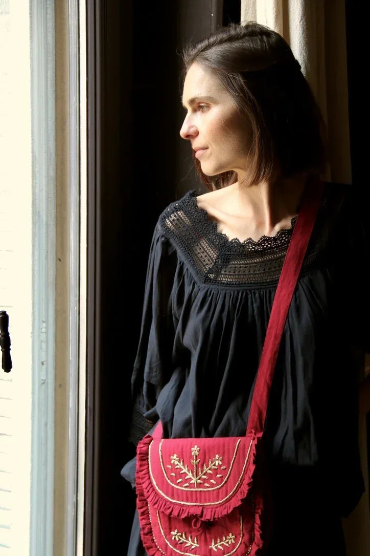 Woman in black dress with lace neckline and red embroidered crossbody bag by window