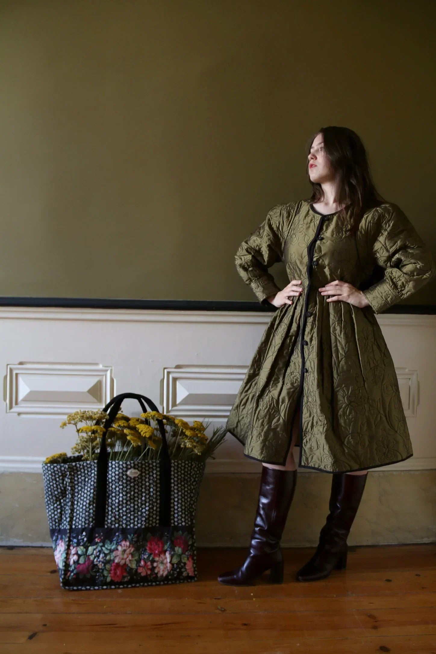 Woman in quilted olive green dress and knee-high brown boots next to floral tote bag with yellow flowers