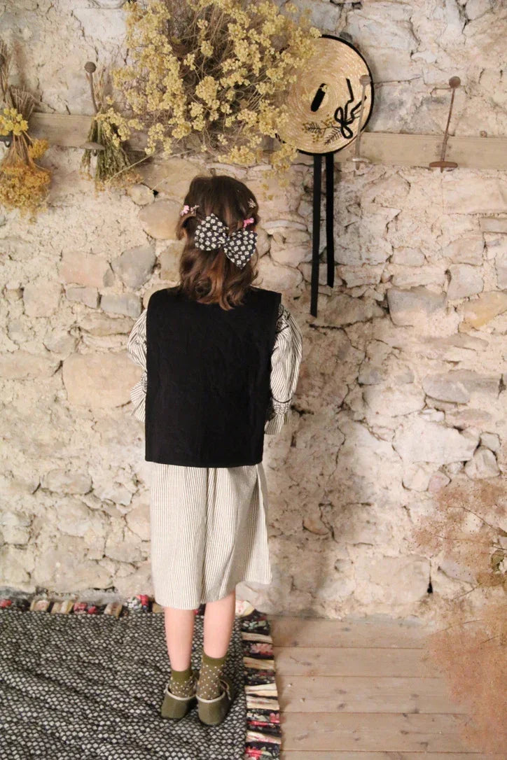 Girl in striped dress and black vest with large bow hair clip, standing by rustic stone wall