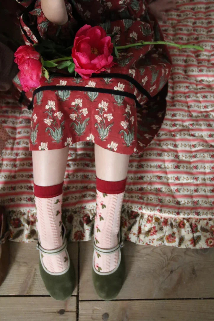 Girl in red floral dress holding bright pink flowers, wearing patterned socks and green shoes, sitting on floral patterned textile.