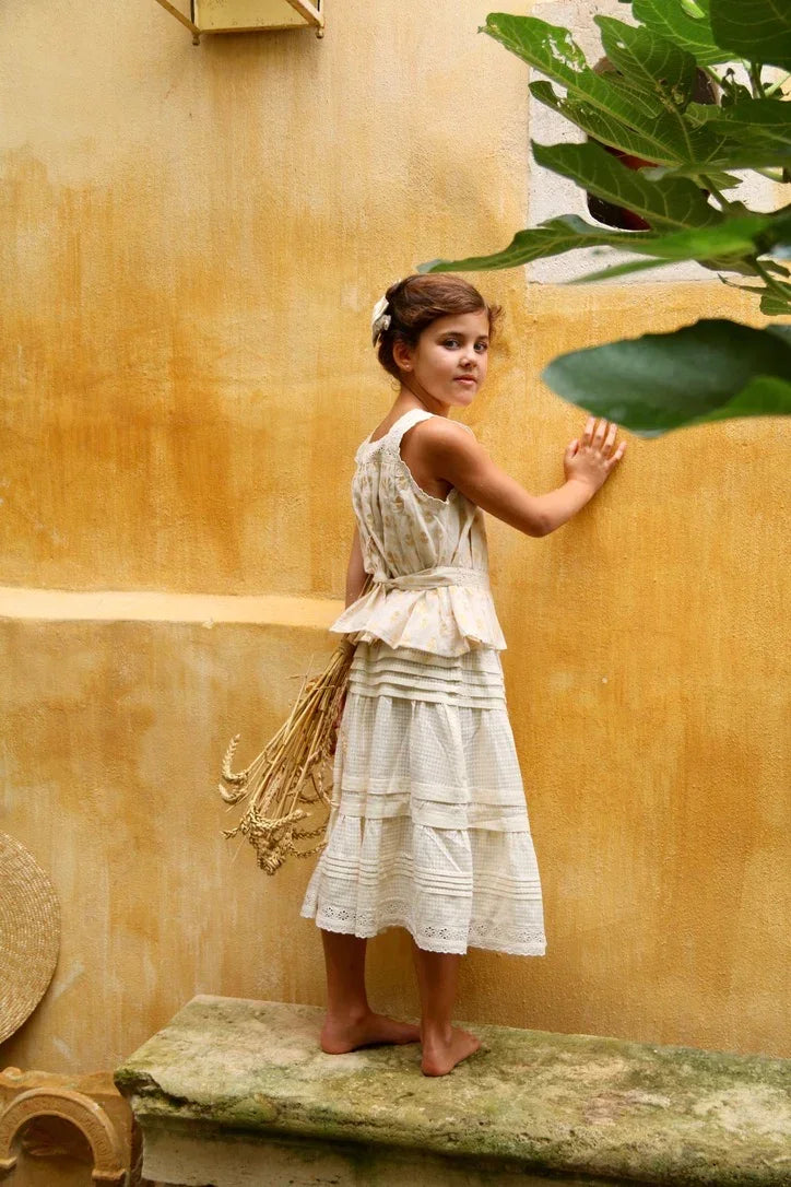 Girl in vintage-style children’s dress with lace details holding dried wheat against yellow wall