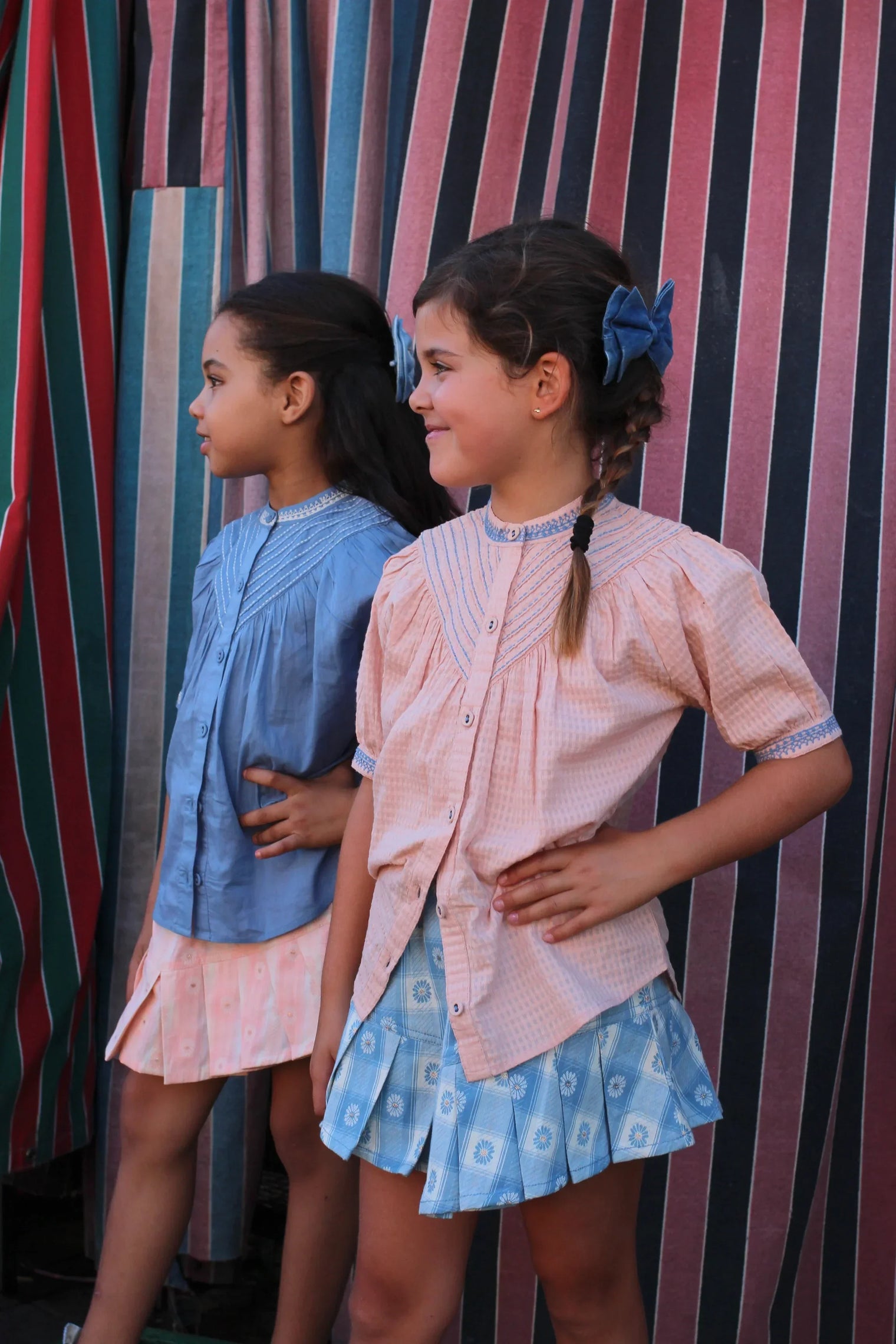 Two girls in pastel blouse and patterned skirt outfits, standing by colorful striped fabric