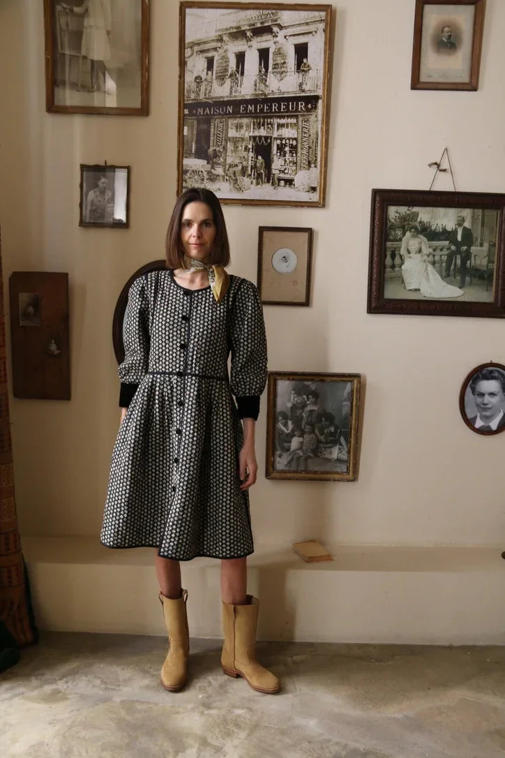 Woman in patterned button-front dress with boots, standing by vintage framed photos on wall