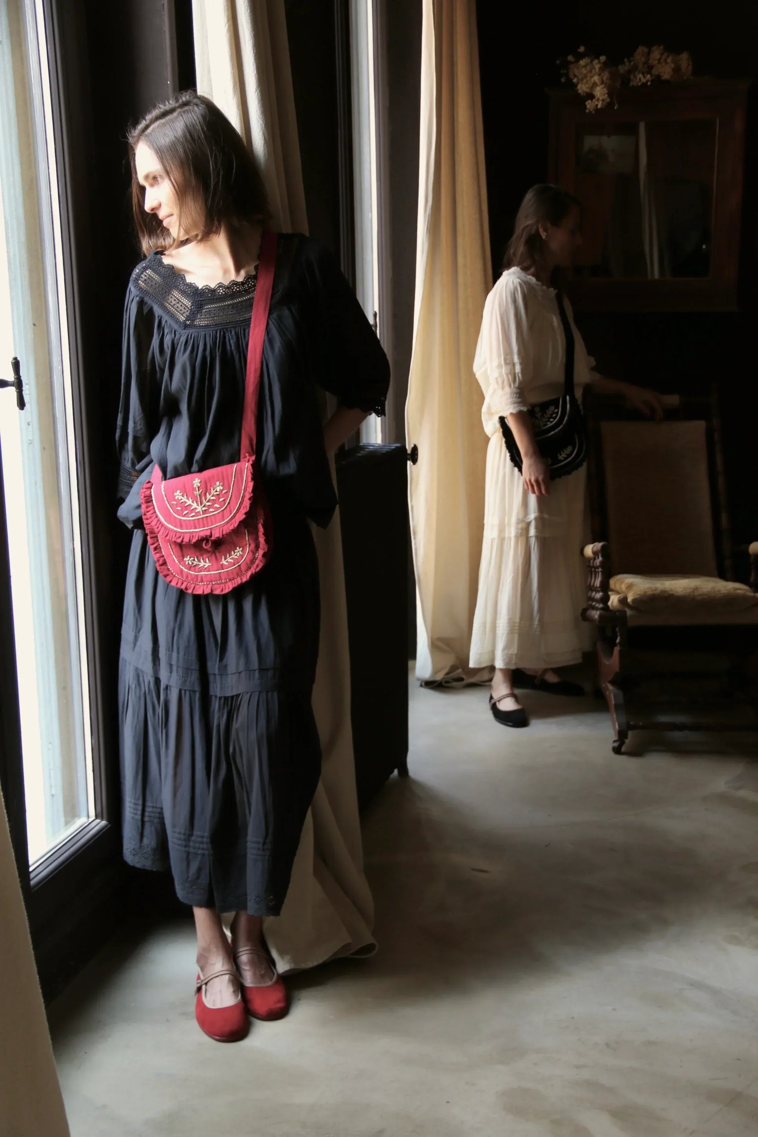 Two women in vintage-inspired dresses with embroidered crossbody bags indoors by a window.