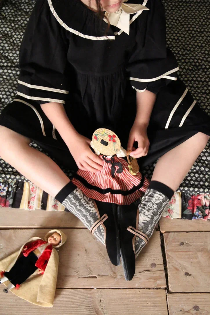 Child in a vintage-inspired black dress with a lace collar, patterned socks, holding a fabric doll