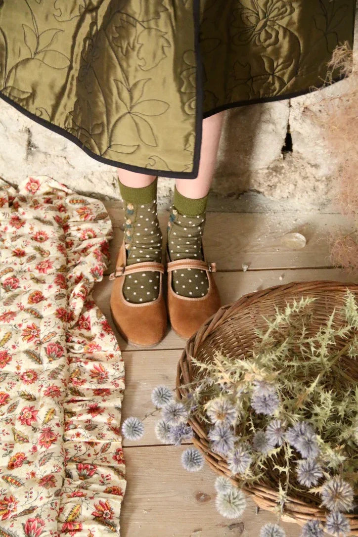 Child in green quilted skirt, polka dot socks, and brown shoes on rustic wood floor with floral textile and wicker basket