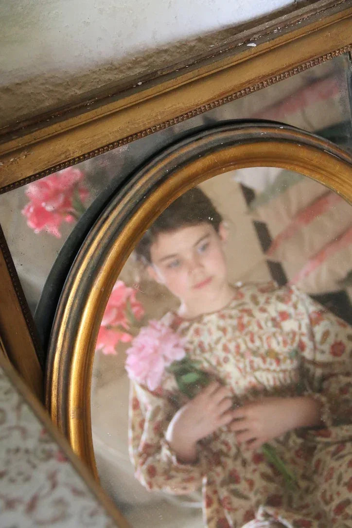Child in vintage floral dress holding pink flowers, reflected in an ornate mirror