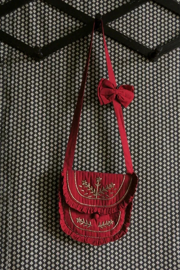Red embroidered children's shoulder bag with ruffles and matching bow on patterned wall