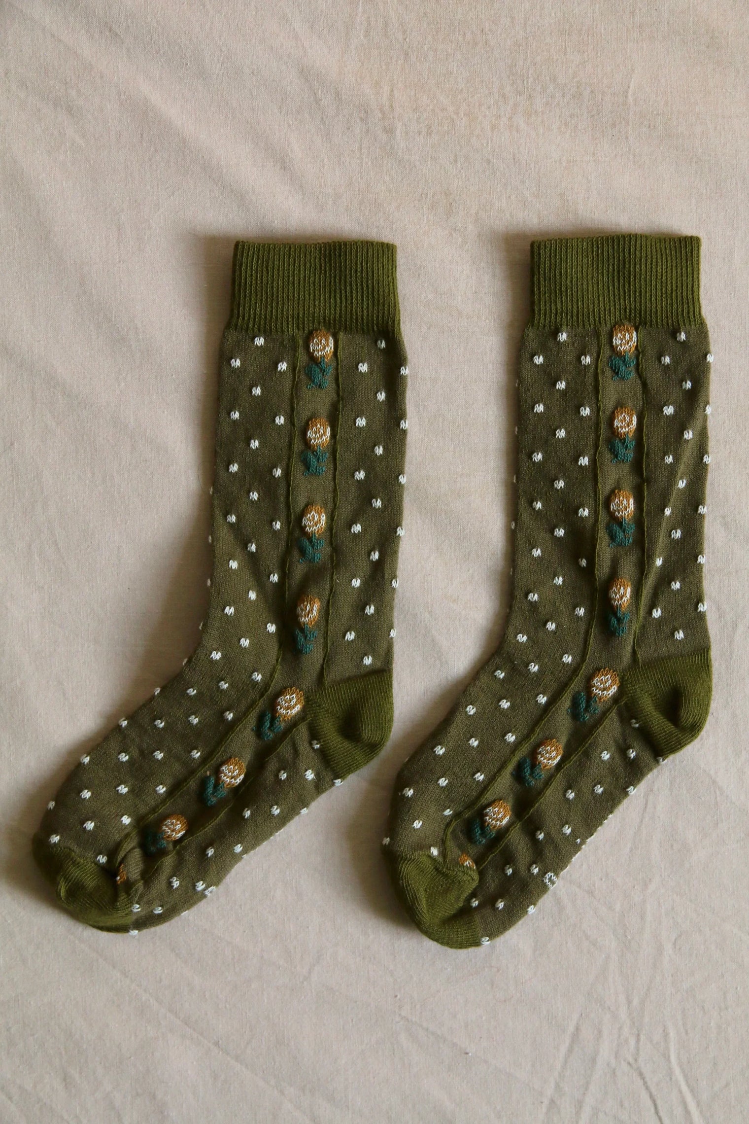 Olive green kids socks with white polka dots and floral embroidery, laid flat on fabric.
