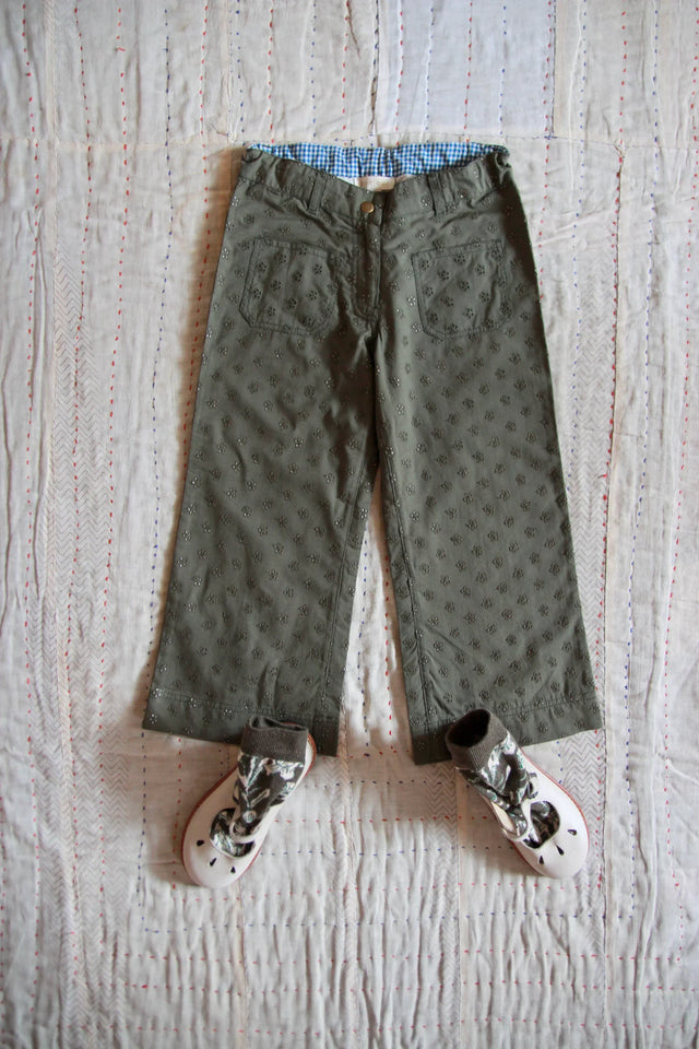 Embroidered green children's pants with front pockets and white shoes on stitched quilt
