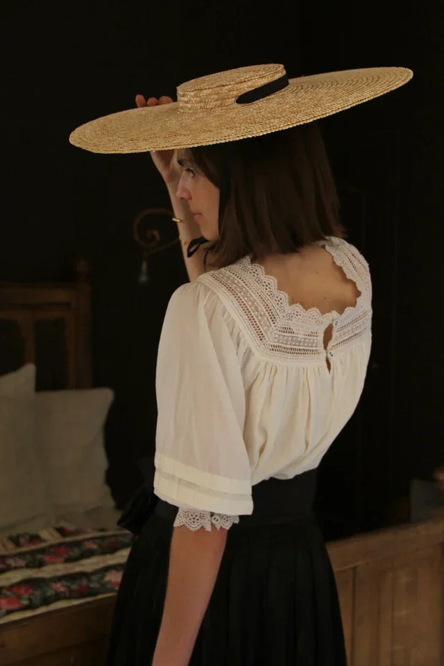 Woman in a vintage lace blouse and wide straw hat standing indoors, classic linen style.