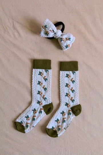 Children’s white knit socks with green accents and floral print, matching bow hair tie on beige background