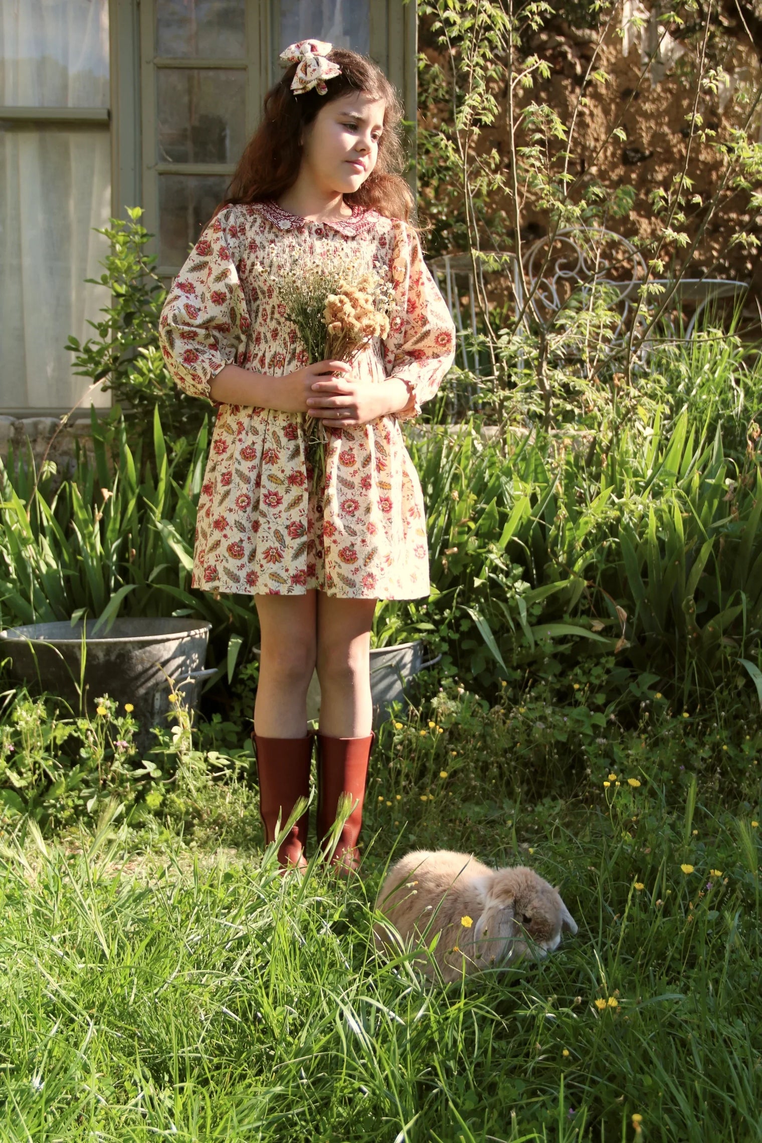 Girl in floral print dress holding bouquet, standing in garden with brown boots and rabbit nearby
