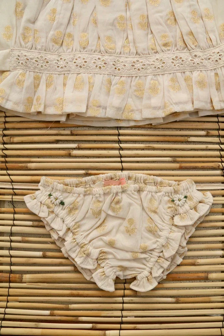 Baby bloomer with gold floral print and ruffle detail on light fabric, laid on bamboo mat