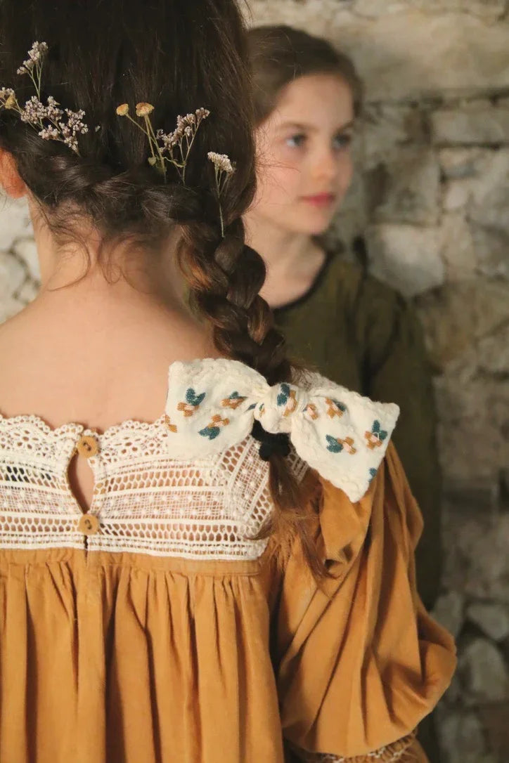 Girl in mustard dress with lace detail, floral hair braid and embroidered bow clip