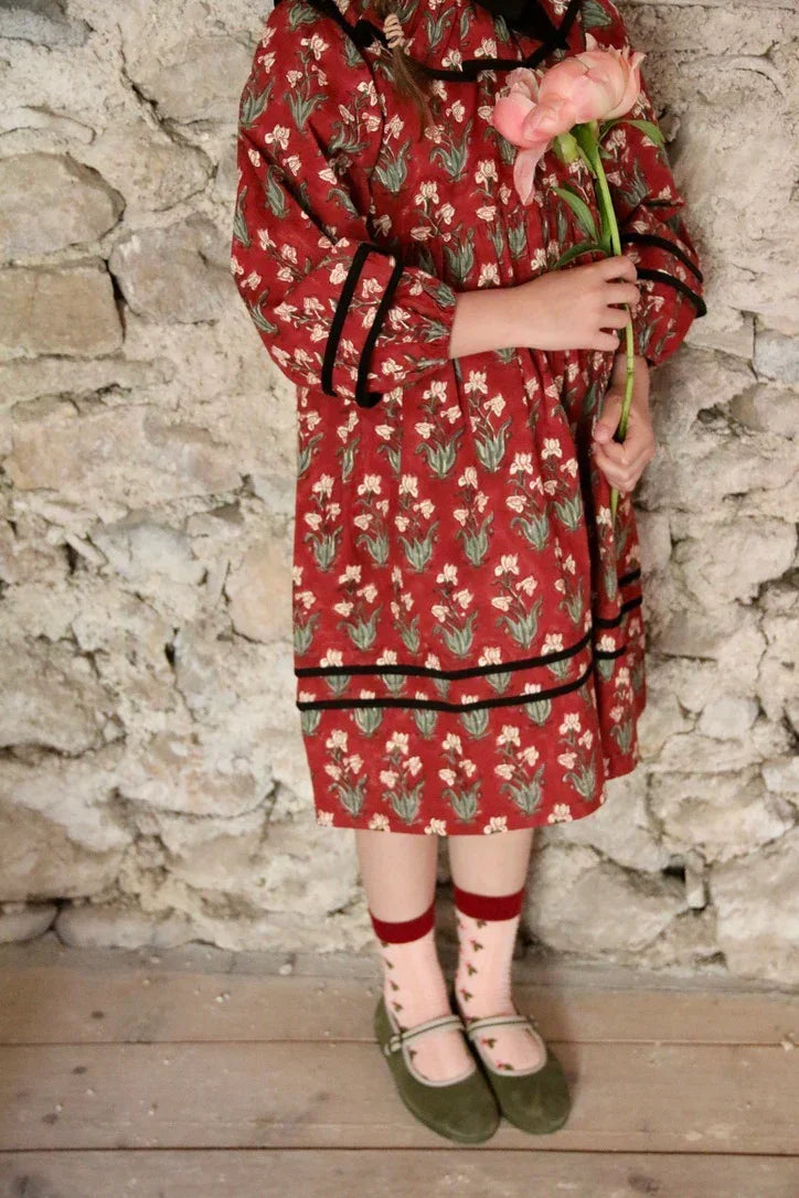 Girl in a red floral dress holding a pink flower, standing by a rustic stone wall