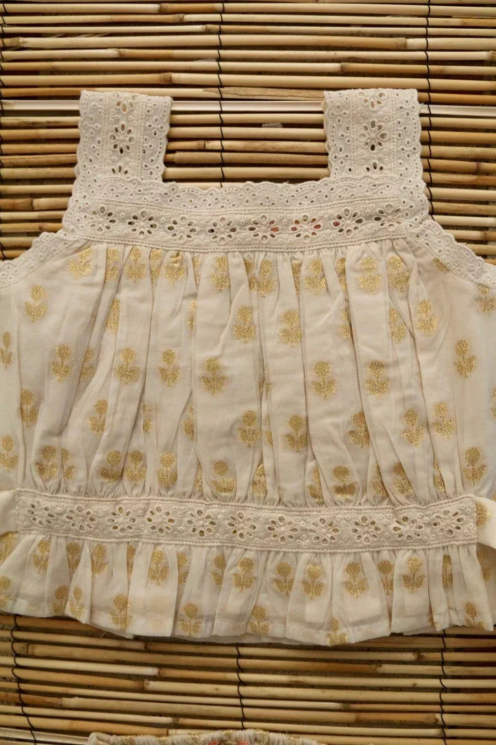 White cotton children's top with gold floral print and lace details, displayed on bamboo backdrop.