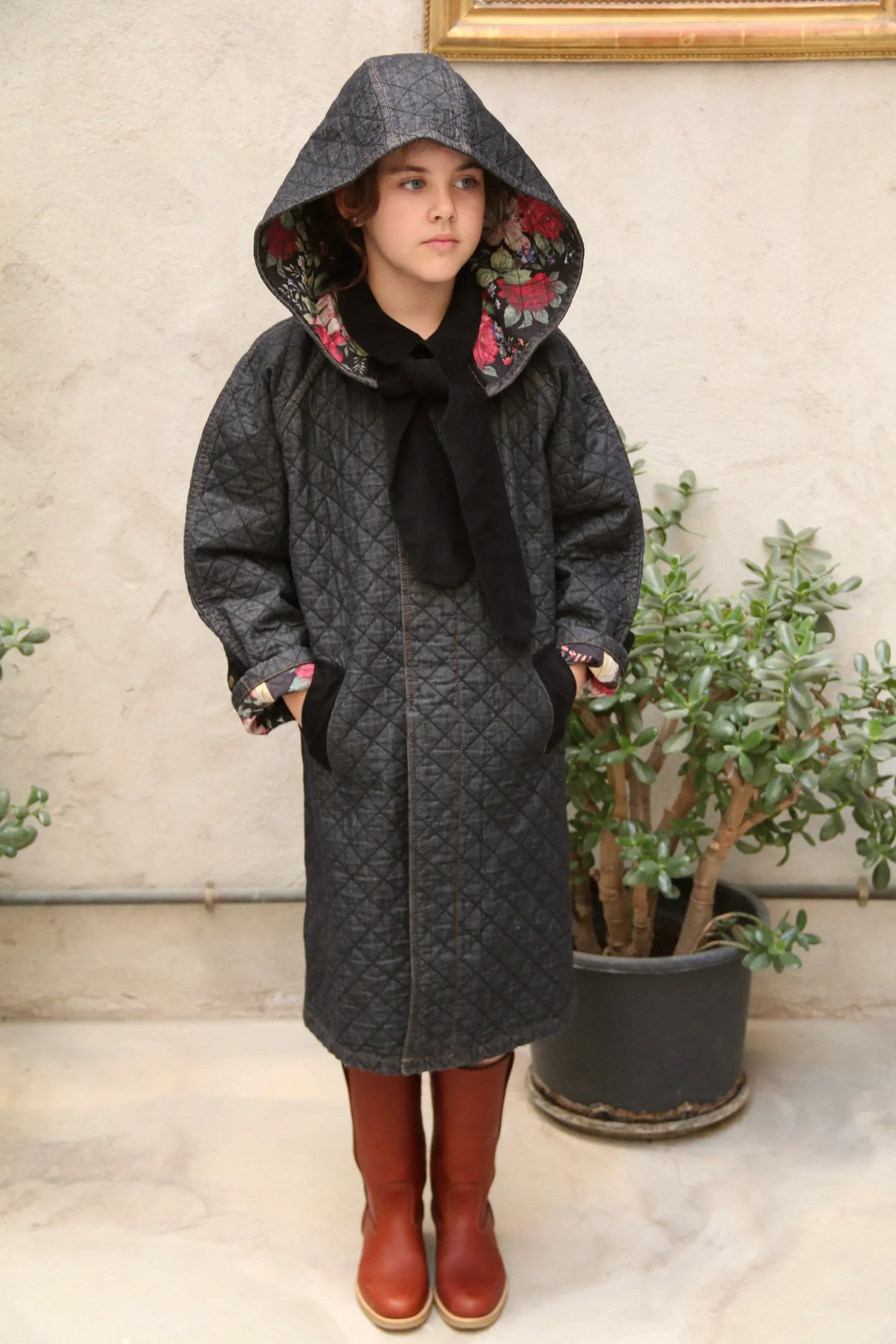 Child in quilted dark grey hooded coat with floral lining and brown boots, indoor setting