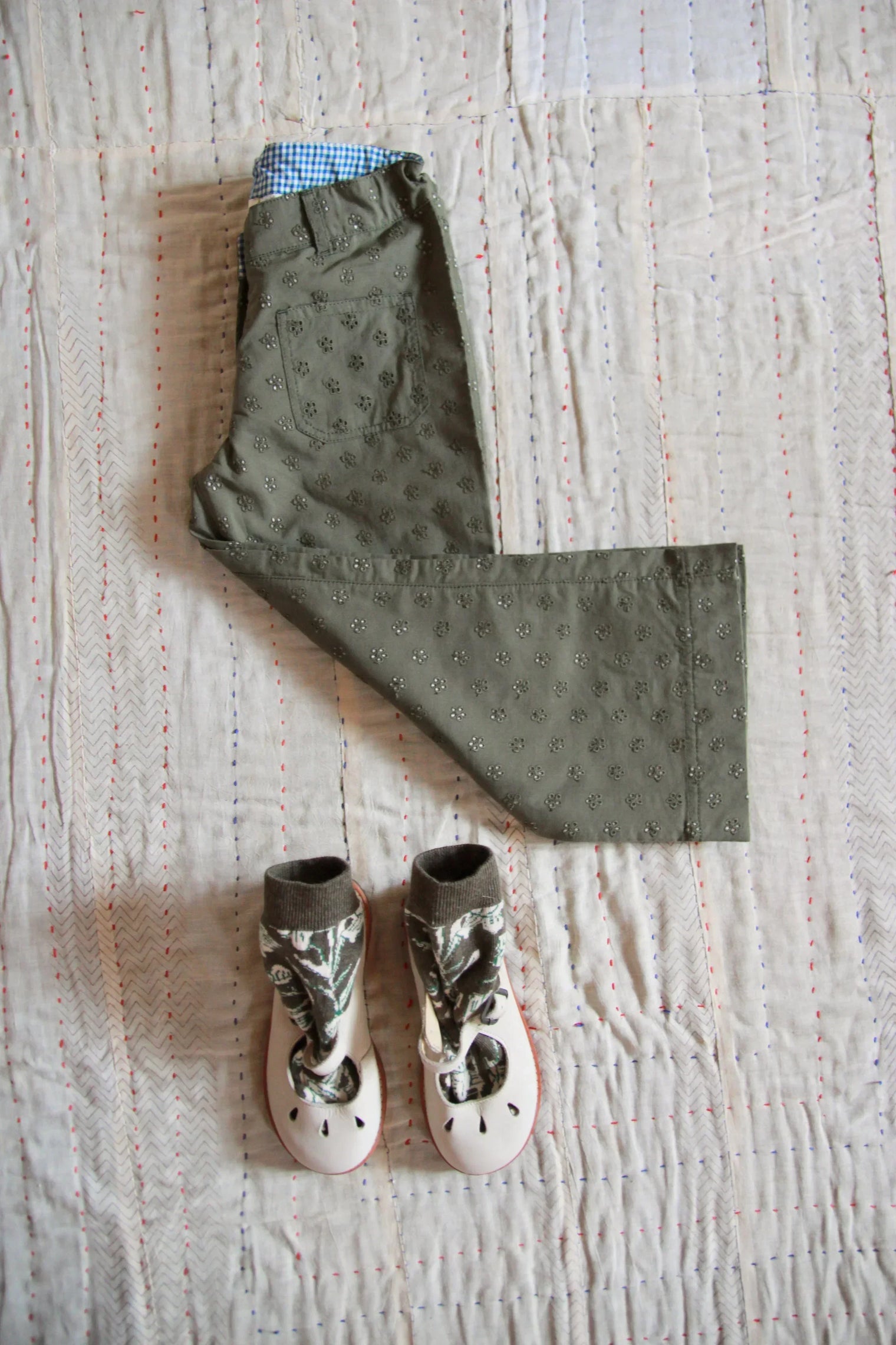 Olive green eyelet embroidered kids pants with blue gingham waistband and floral baby shoes on quilted linen background