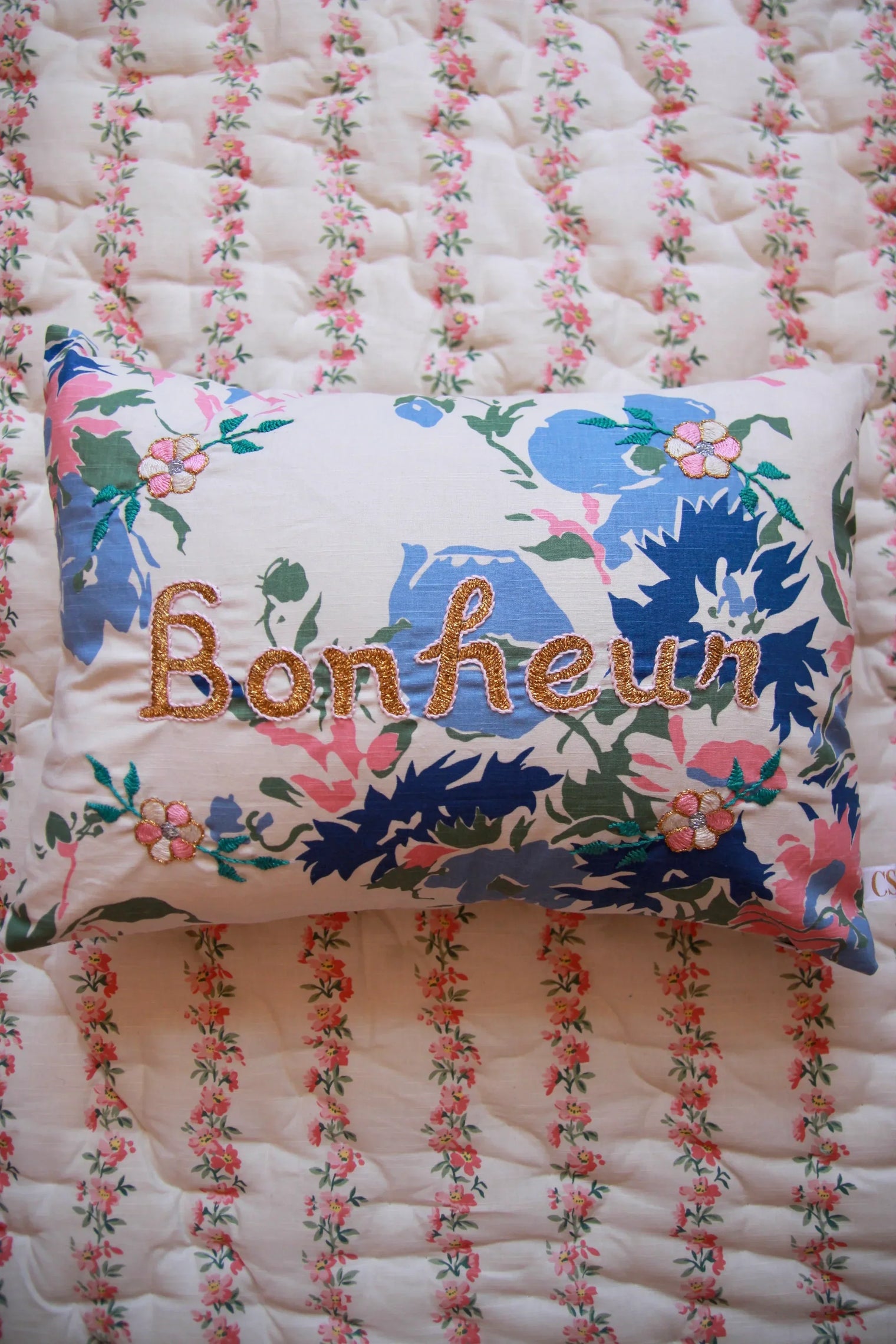 Floral embroidered pillow with 'Bonheur' text on a quilted floral bedspread, kids linen
