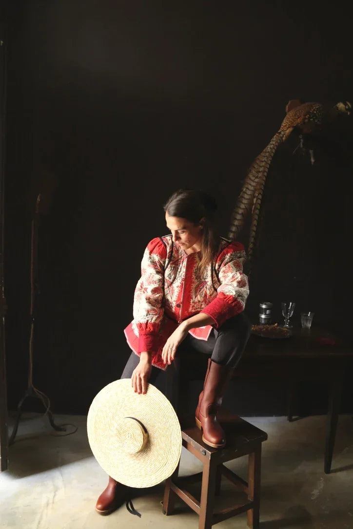 Woman in floral embroidered jacket and tall boots holding a large straw hat indoors
